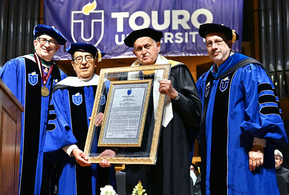 Touro President Dr. Alan Kadish, Chairman of the Board Zvi Ryzman, Dean Robert Goldschmidt, and Executive Vice President Rabbi Moshe Krupka stand in regalia on stage with a Touro University sign in the background. Dean Robert Goldschmidt holds the framed honorary doctorate that was presented to Zvi Ryzman.