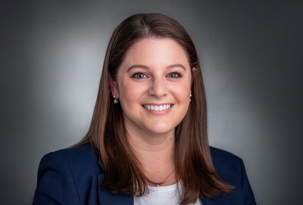 Headshot of Emily Messing, a woman with long brown hair, wearing a white crew neck shirt and navy blue blazer