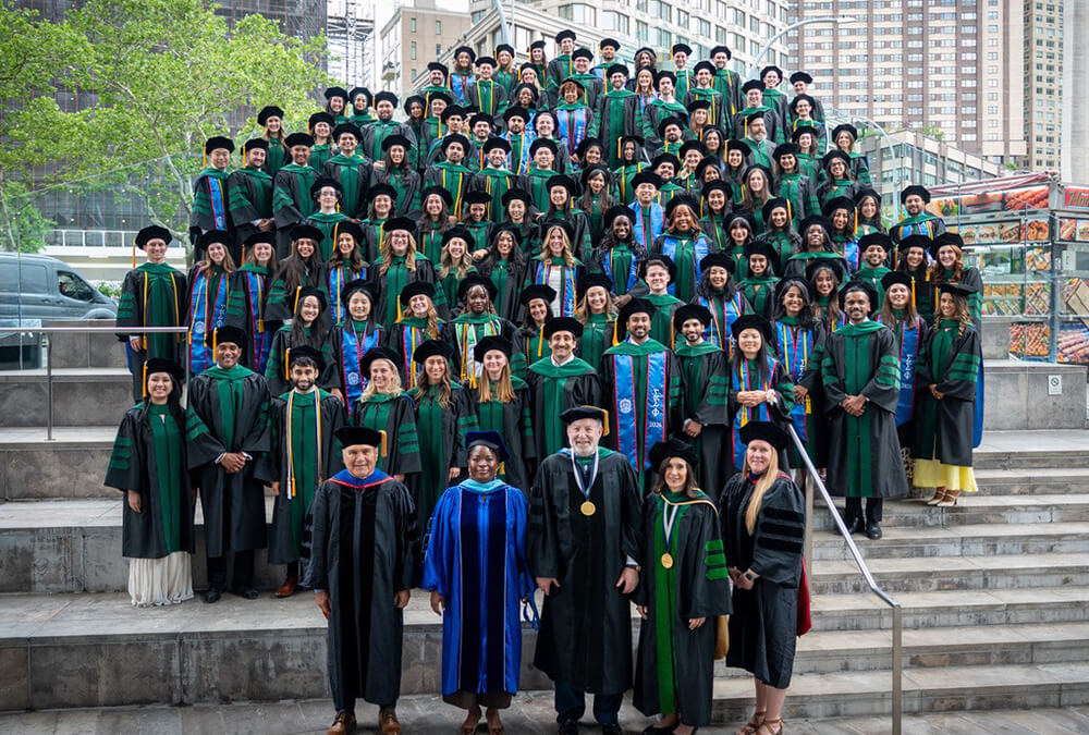 Over 100 young men and women standing on steps together lined up outside at Lincoln Center plaza smiling in caps and gowns. 