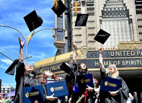 four young women graduates standing in street outside theater after commencement ceremonies tossing their caps in the air against the blue sky. 