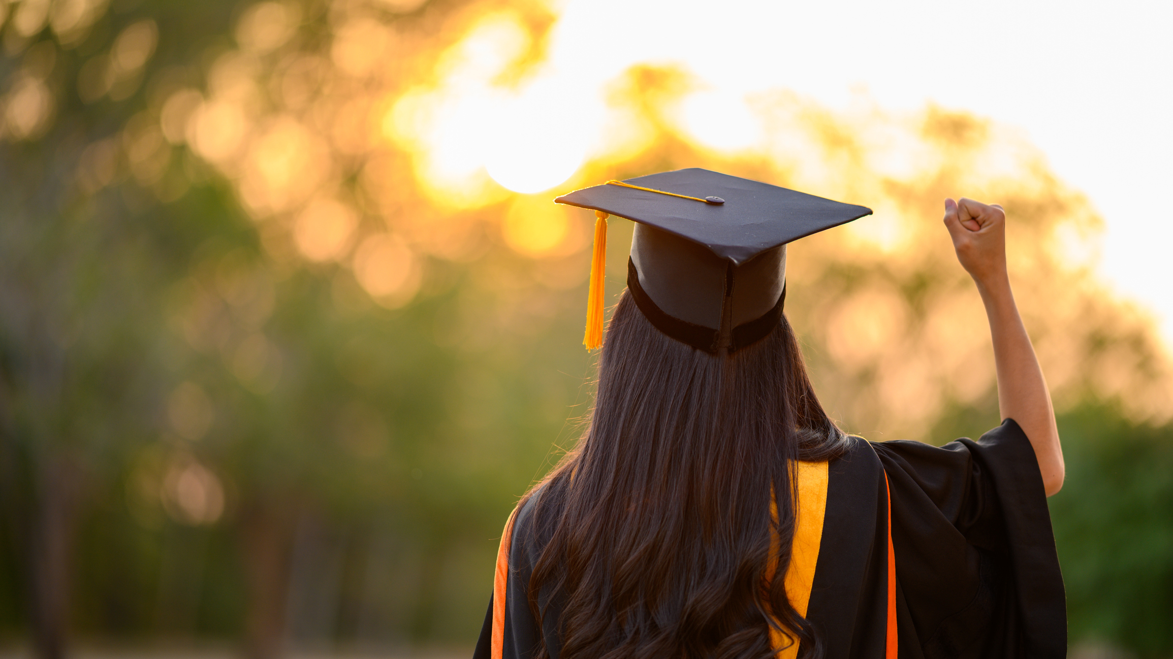 Back of female graduate wearing black cap & gown with yellow tassel waiting outside to attend the commencement ceremony at the university. Afternoon sun and trees blurred in the background.